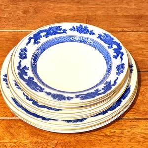 May include: A stack of white ceramic plates featuring a blue dragon pattern along the rim. Each plate has a gold-trimmed edge and a central white circular area. The blue design includes intricate dragon motifs and floral elements on the border.