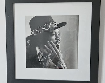 May include: Black and white framed portrait of a man wearing a hat with circular embellishments. The man is looking to the side, with his hand near his face. The portrait is in a black frame.