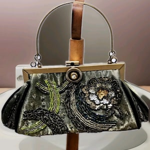 May include: An ornate evening bag in a muted green colour, embellished with detailed beadwork and sequins. The bag showcases a floral design and a metal frame with a handle. The bag is displayed on a stand.