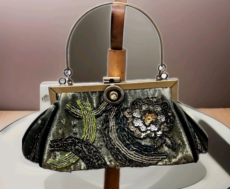 May include: An ornate evening bag in a muted green colour, embellished with detailed beadwork and sequins. The bag showcases a floral design and a metal frame with a handle. The bag is displayed on a stand.