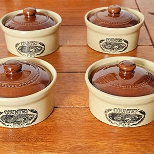 May include: A set of four round ceramic pots with lids. Each pot features a cream-colored base with a dark brown glazed lid and a black printed graphic on the side that reads "COUNTRY POT" with an illustration of vegetables.