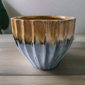 May include: A ceramic planter with a ribbed design. The top section is a warm brown colour that fades into a cool grey-blue at the bottom. The planter has a wide opening and sits on a wooden surface.