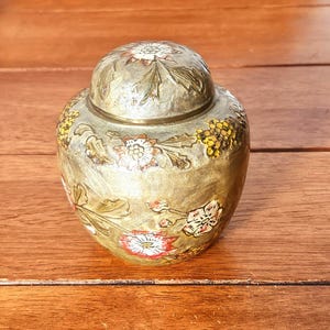 May include: A small, round, metallic jar with a matching lid, featuring an embossed and hand-painted floral design. The surface has a muted gold finish with detailed flowers in shades of red, white, and yellow, accented by green leaves.