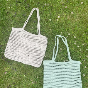 May include: Two crocheted tote bags, one in a light beige color and one in a light green color, are lying on a bed of green grass. The bags have a simple, open weave pattern.