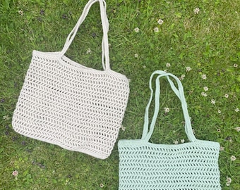 May include: Two crocheted tote bags, one in a light beige color and one in a light green color, are lying on a bed of green grass. The bags have a simple, open weave pattern.
