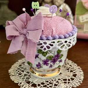 Puede incluir: Un alfiletero de color morado y blanco con forma de cupcake, con un borde de encaje y un lazo morado. El alfiletero está sobre un encaje de ganchillo blanco.
