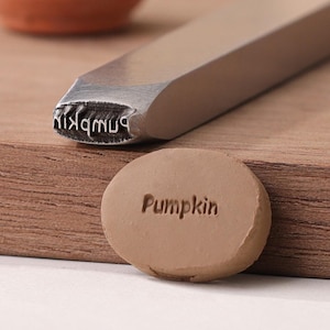 Peut inclure: Un tampon en métal avec le mot "Pumpkin" gravé dessus. Le tampon est utilisé pour créer une empreinte dans l'argile. L'argile est de couleur brun clair et porte l'inscription "Pumpkin".
