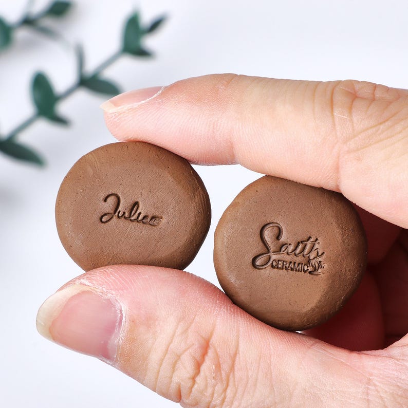 May include: Two small, round, brown clay discs with stamped text. The left disc features the name Julie in cursive script, while the right disc displays the text Sait's CERAMIC with a small graphic icon of a plant or tree underneath the lettering.