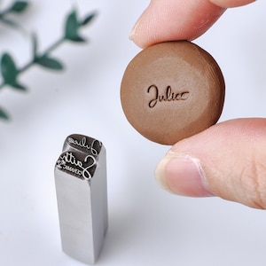 May include: A silver metal stamp with a rectangular base and a custom engraved design featuring the text "Juliee" and "Faith CERAMIC" with a palm tree icon, alongside a small brown circular clay disc imprinted with the name "Juliee" in script.