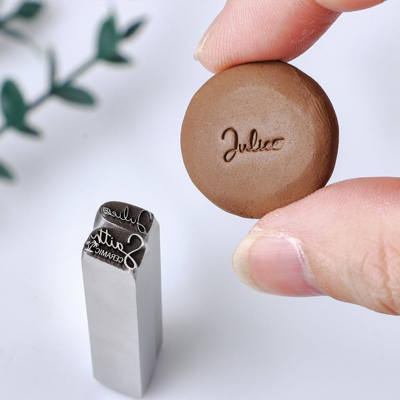 May include: A silver metal stamp with a rectangular base and a custom engraved design featuring the text "Juliee" and "Faith CERAMIC" with a palm tree icon, alongside a small brown circular clay disc imprinted with the name "Juliee" in script.