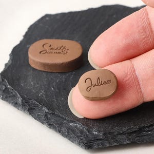 May include: Two small, oval-shaped brown clay stamps. One stamp is held, displaying the name Julie with a small leaf icon engraved on the surface. The second stamp rests nearby, featuring the text Sattie Ceramic engraved on its flat brown surface.