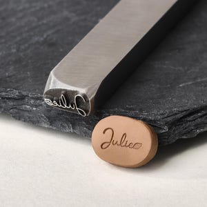 May include: A silver-toned metal stamping tool with a rectangular shaft and a custom head featuring the name Julie in cursive script followed by a small leaf icon, alongside a tan oval clay piece embossed with the same name and leaf design.