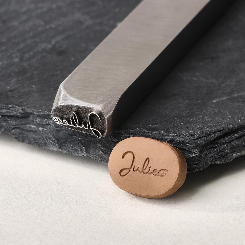 May include: A silver-toned metal stamping tool with a rectangular shaft and a custom head featuring the name Julie in cursive script followed by a small leaf icon, alongside a tan oval clay piece embossed with the same name and leaf design.