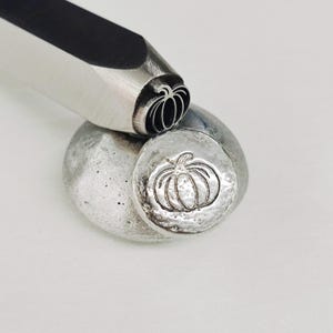 May include: A metal stamp with a pumpkin design is being used to create an impression on a piece of metal.