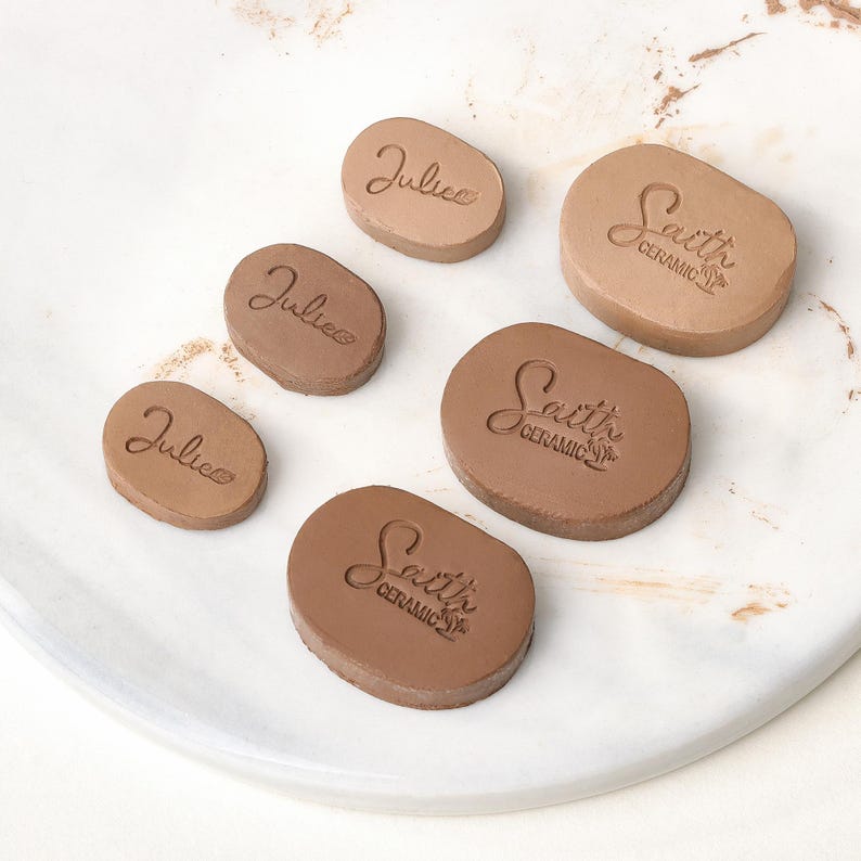 May include: Six oval-shaped clay stamps in varying sizes, featuring embossed text. Three stamps display the name Julie, while the other three larger stamps feature the text Saith Ceramic with a small palm tree icon, all in a light brown tone.