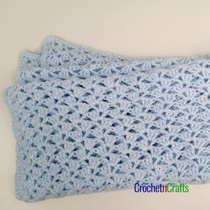 Puede incluir: Una manta de bebé de ganchillo de color azul claro con un delicado patrón de encaje. La manta está doblada por la mitad y está sobre una superficie blanca.