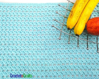 Op de afbeelding: Een lichtblauw gehaakt placemat met een geweven patroon. De placemat ligt op een wit oppervlak met een metalen fruitmand met bananen en een appel.
