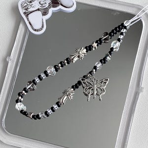 May include: A beaded phone charm with black beads, clear beads, and silver accents. The charm features a silver butterfly pendant, cross charms, and a small sticker of a video game controller.
