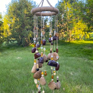 May include: A handcrafted wind chime with a jute-wrapped ring and hanging strands. The strands feature various natural elements like wooden beads, seed pods, and dark brown, teardrop-shaped pieces. The wind chime is suspended against a backdrop of green trees and grass.