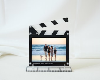 May include: A black and white clapperboard-shaped photo frame with a family photo inside. The clapperboard has a black frame with a white and black striped top. The photo shows a family walking on a beach. The frame is on a clear stand.