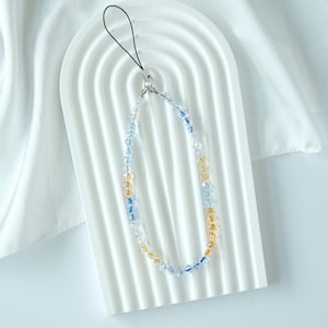 May include: A beaded phone charm featuring clear, blue, and yellow beads. The charm is attached to a black loop and is displayed on a white, arched surface. The background is white, draped fabric.