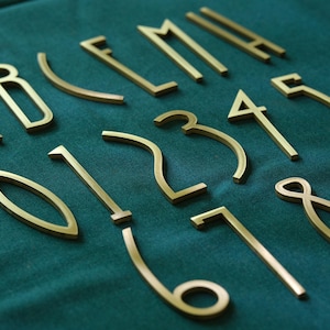 K&ouml;nnte beinhalten: Goldfarbene Metallbuchstaben und -zahlen auf einem t&uuml;rkisgr&uuml;nen Stoff angeordnet. Das Set enth&auml;lt die Buchstaben A, B, E, L, M und die Zahlen 0, 1, 2, 3, 4, 5, 6, 7, 8 und 9. Die Buchstaben und Zahlen sind in einer serifenlosen Schriftart.