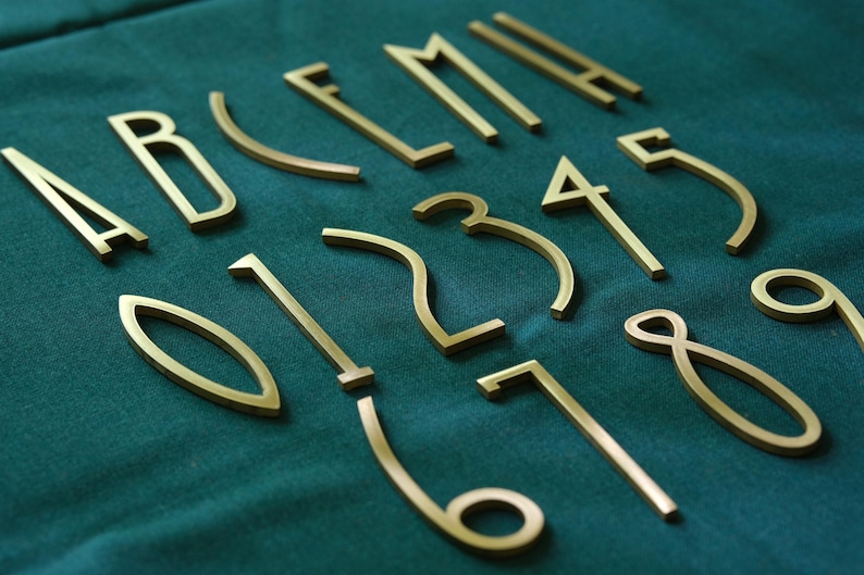 K&ouml;nnte beinhalten: Goldfarbene Metallbuchstaben und -zahlen auf einem t&uuml;rkisgr&uuml;nen Stoff angeordnet. Das Set enth&auml;lt die Buchstaben A, B, E, L, M und die Zahlen 0, 1, 2, 3, 4, 5, 6, 7, 8 und 9. Die Buchstaben und Zahlen sind in einer serifenlosen Schriftart.