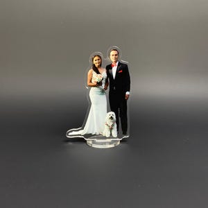 Peut inclure: Un cake topper de mariage en acrylique transparent représentant une mariée en robe blanche et un marié en costume noir, avec un petit chien blanc. La mariée tient un bouquet. Les personnages sont sur une base transparente.