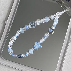May include: A beaded phone charm featuring a mix of clear, white, and light blue beads. The charm includes star and heart-shaped beads. The charm is displayed on a reflective surface.