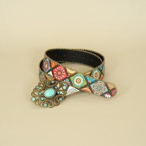 May include: A coiled belt featuring a repeating diamond pattern in vibrant colours including red, orange, blue, and white. The belt has a decorative antique brass-toned buckle with intricate scrollwork and a central oval turquoise-coloured stone.