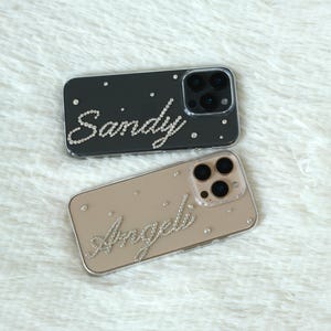 Könnte beinhalten: Zwei Handyhüllen, eine schwarze und eine beige, werden gezeigt. Die schwarze Hülle hat den Namen "Sandy" in Strasssteinen, und die beige Hülle hat den Namen "Angel" in Strasssteinen. Beide Hüllen haben einen transparenten Rand.