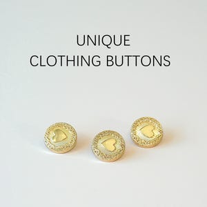 May include: Three circular gold-toned buttons featuring a central heart motif surrounded by a textured border. The text UNIQUE CLOTHING BUTTONS is printed above the buttons. Each button has a smooth, polished finish on the heart and a detailed rim.