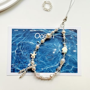 May include: A beaded phone charm with pearls, clear beads, and seashell charms. The charm is silver and white, with a starfish and a ring. The background features a blue ocean image with the word "Ocean".