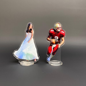 Könnte beinhalten: Zwei Acrylfiguren: eine Frau in einem weißen und blauen Kleid und ein Footballspieler in roter Uniform. Das Kleid der Frau hat einen fließenden Rock mit blauen und grünen Farbtönen. Der Footballspieler hält einen Football und trägt einen Helm.