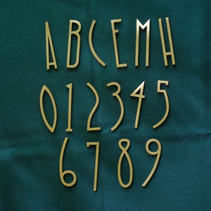 May include: Gold-colored metal letters and numbers arranged on a dark teal fabric. The letters A, B, C, E, M, and H are visible, along with the numbers 0 through 9. The font is a thin, elegant sans-serif style.