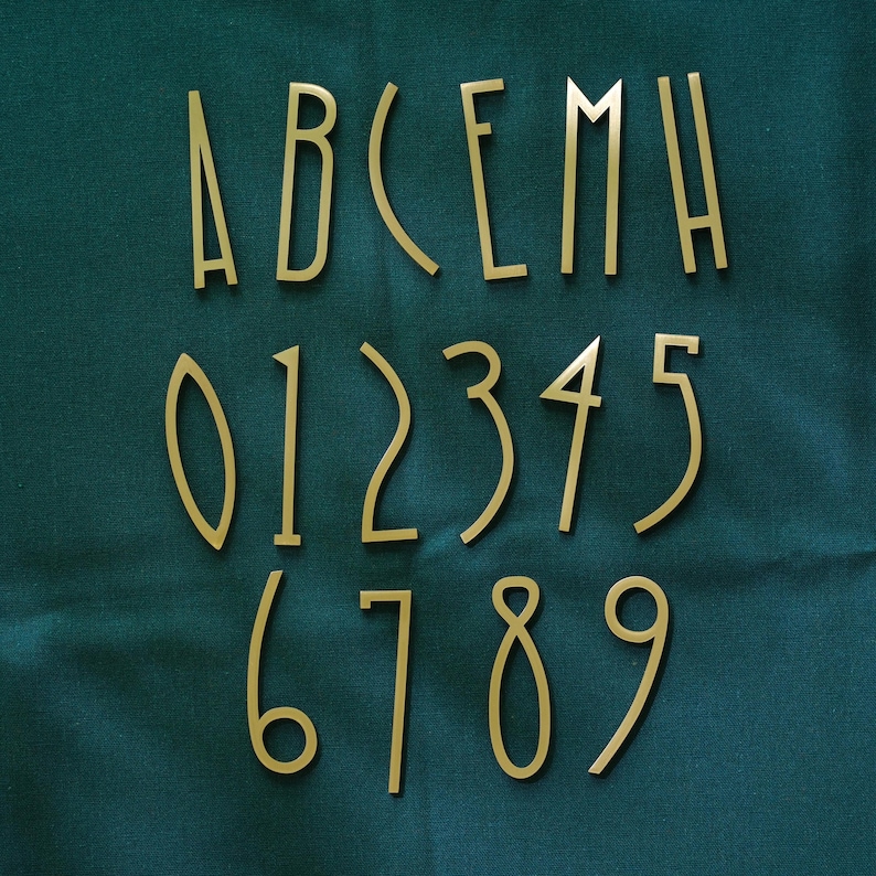 K&ouml;nnte beinhalten: Goldfarbene Metallbuchstaben und -zahlen auf einem dunkelblauen Stoff angeordnet. Die Buchstaben A, B, C, E, M und H sind sichtbar, ebenso wie die Zahlen 0 bis 9. Die Schriftart ist ein d&uuml;nner, eleganter Sans-Serif-Stil.
