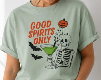 May include: A light sage green t-shirt featuring a graphic of a skeleton holding a martini glass with a pumpkin and ghost garnish. The text reads "GOOD SPIRITS ONLY" in orange letters. Two black bats are printed to the left of the graphic.