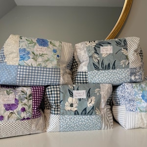 May include: A collection of handmade fabric pouches with floral and patterned designs. Each pouch is made from a patchwork of fabrics in various colours, including blue, white, purple, and green. The pouches have a label that reads "Stitch By Adria".
