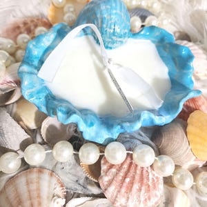 May include: A blue seashell candle holder with a white candle inside. The holder is surrounded by white pearls and seashells.
