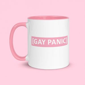 May include: White ceramic mug with a pink interior, handle, and a pink rectangular text box that reads "[GAY PANIC]" in white letters. The mug is set against a solid pink background.