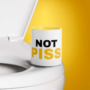 Puede incluir: Una taza de cerámica blanca con un borde amarillo y el texto "NOT PISS" en letras negras y amarillas. La taza está sentada en un asiento de inodoro blanco.