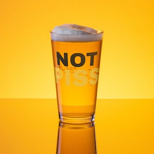 May include: A tall glass filled with a light yellow beer with a white head. The glass has the words "NOT PISS" printed on it.