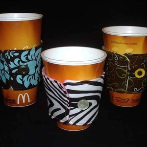 May include: Three coffee cups with fabric sleeves. The sleeves are different patterns: blue and white floral, black and white zebra print, and brown with swirls.