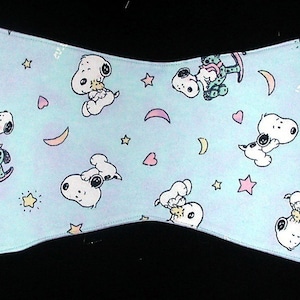 Könnte beinhalten: Ein hellblauer Spucktuch für Babys mit weißem Rand. Das Tuch hat ein sich wiederholendes Muster aus einem Cartoon-Hund, der ein Schaukelpferd reitet, Sternen, Monden und Herzen. Der Text "My Little Snoopy" ist auf das Tuch gedruckt.