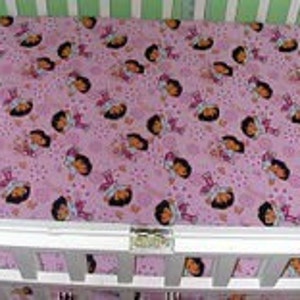 Puede incluir: Una sábana de cuna rosa y morada con un patrón repetido de una niña de dibujos animados con cabello castaño y un vestido rosa. La niña está rodeada de corazones y estrellas rosas.