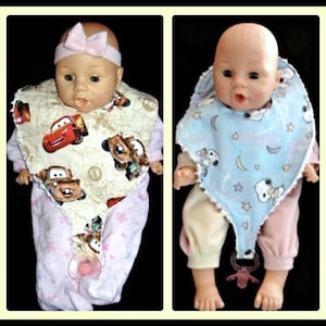 May include: Two baby dolls wearing bibs. The doll on the left is wearing a pink outfit with a Cars-themed bib. The doll on the right is wearing a yellow and pink outfit with a blue bib featuring a Snoopy design.