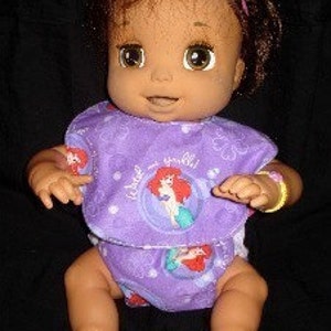 Pode incluir: Uma boneca de bebê de pele morena usando um babador roxo e branco com uma imagem de desenho animado de Ariel da Pequena Sereia. O babador diz "Little Mermaid". A boneca está usando uma fralda roxa e branca com a mesma imagem de Ariel. A boneca tem cabelo castanho e olhos castanhos.