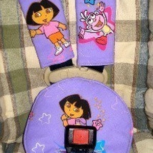 May include: Purple Dora the Explorer themed car seat belt covers and a matching booster seat cushion. The covers feature Dora and Boots with stars. The cushion has a picture of Dora and the words "Let's Go!"
