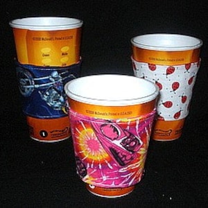 May include: Three white coffee cups with colourful fabric sleeves. The sleeves have different designs, including a motorcycle, ladybirds, and a pink and orange design with a rocket ship.