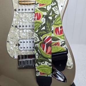 May include: A guitar strap with a botanical design featuring green and red pitcher plants on a cream background. The strap is attached to a metallic gold electric guitar with a pearlescent pickguard.